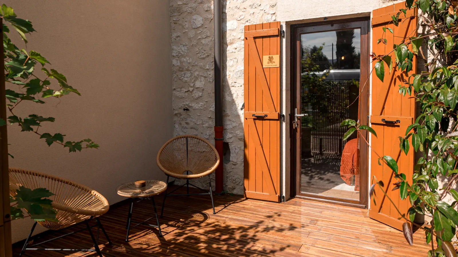 Petit espace extérieur avec deux chaises en rotin, une table basse et une porte en bois orange ouverte sur un jardin