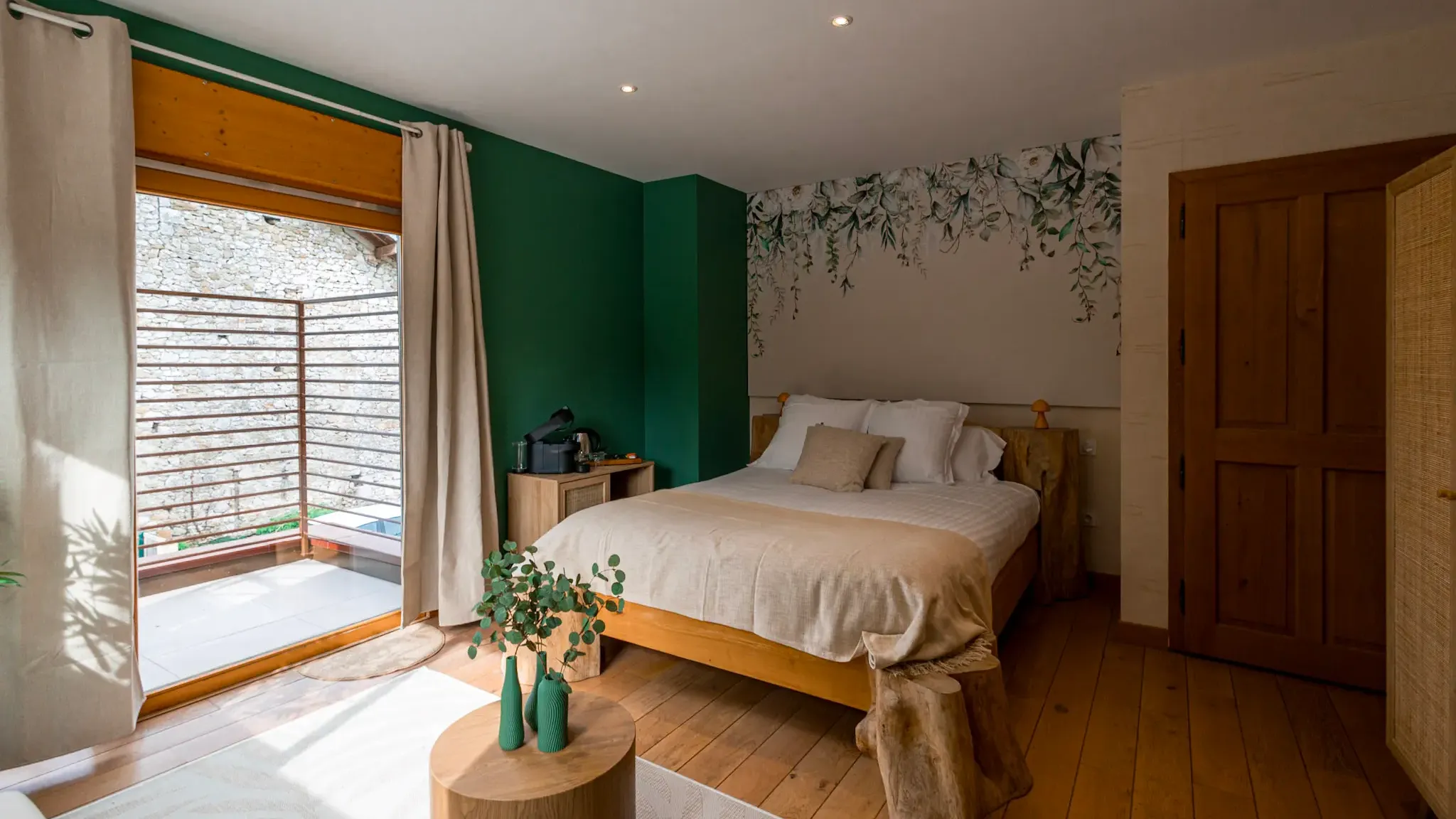 Chambre avec un lit en bois naturel, une fenêtre ouverte avec des rideaux, un mur vert et une décoration murale florale, un petit meuble de chevet et une table basse avec des vases